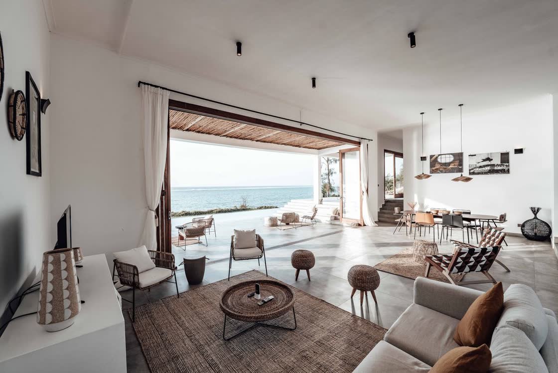 An open-plan living room looking out through a wide opening to the sea, with the camera path drawn across it as a thin orange line that stops before the threshold.